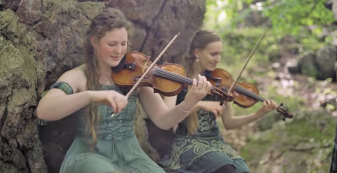 Two fiddlers playing traditional music outdoors beside a stone wall in a wooded setting.