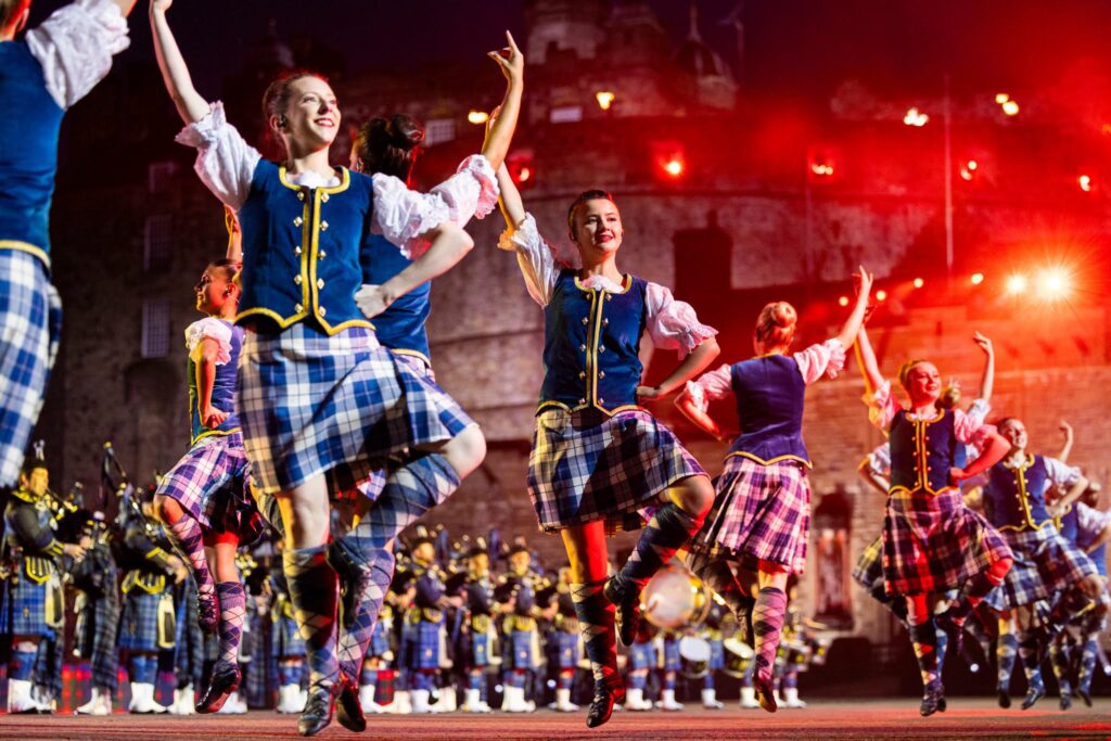 Highland dancers in tartan kilts perform with raised arms as a pipe band plays behind them at the Royal Edinburgh Military Tattoo.