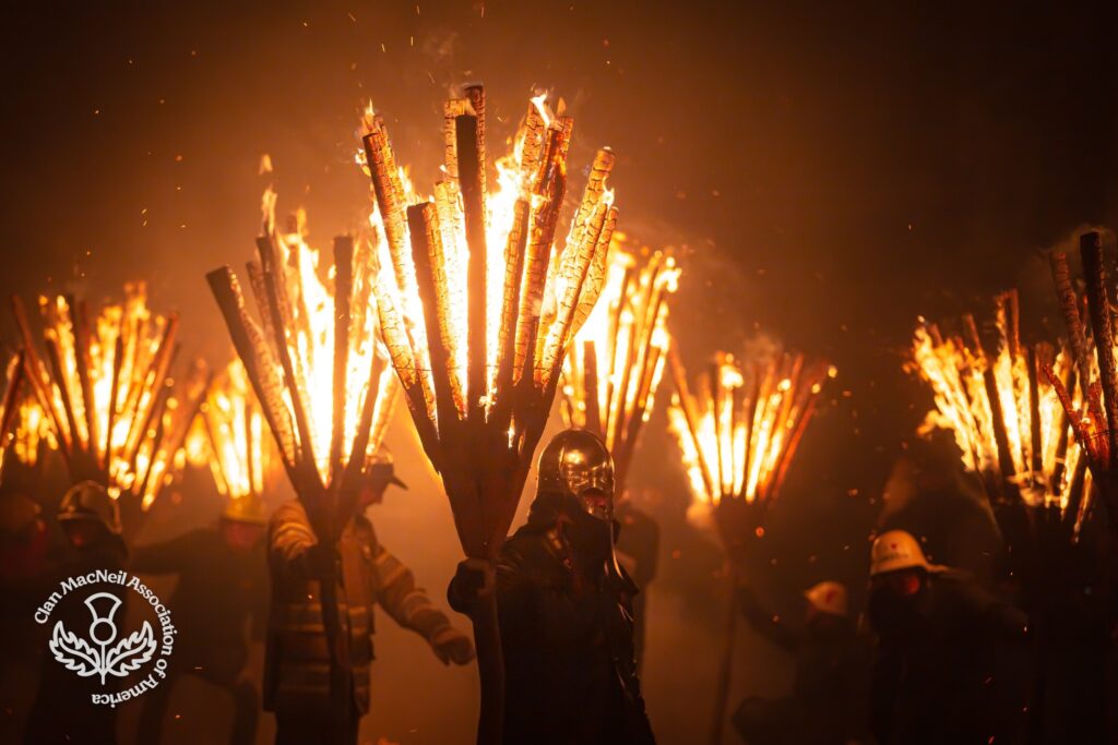 Torchbearers carry huge bundles of burning wood through the night, sparks and smoke filling the air, one figure in front wearing a metal mask.